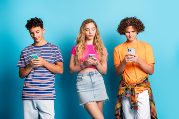 Three friends in bright casual outfits text each other while posing against a blue backdrop in a trendy teen lifestyle stock photo