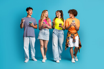 Obraz premium Playful group of friends chatting and texting on phones in bright casual outfits against a vibrant blue backdrop