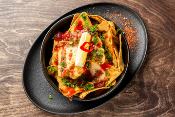 Loaded nachos with melted cheese, ground meat, sour cream, chili, parsley, and sauce served in a black dish. Tasty snack or appetizer in a rustic restaurant setting.