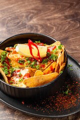Loaded nachos with melted cheese, ground meat, sour cream, chili, parsley, and sauce served in a black dish. Tasty snack or appetizer in a rustic restaurant setting.