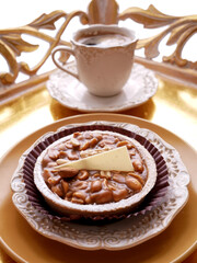A close-up shot shows a small tart topped with nuts and a slice of white chocolate, and a cup of coffee on a decorative tray. The composition suggests a dessert or breakfast setup.