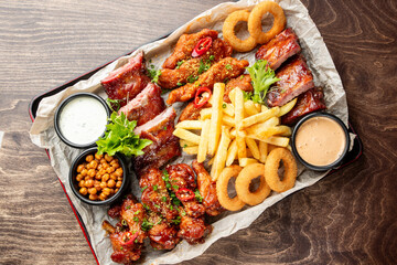 Assorted fried and grilled foods on parchment-lined tray: ribs, wings, fries, onion rings, sauces, chickpeas, herbs. Casual comfort food platter for sharing.