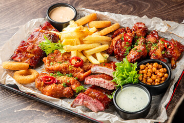 Assorted fried and grilled foods on parchment-lined tray: ribs, wings, fries, onion rings, sauces, chickpeas, herbs. Casual comfort food platter for sharing.