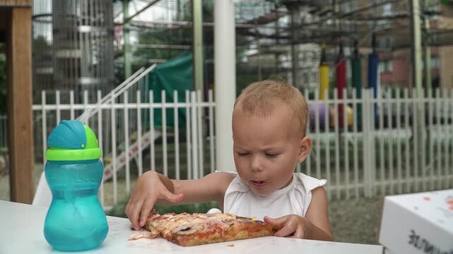 A girl with short hair bites into a pizza, tearing off a large piece with both hands. She eats with concentration, chews, and looks at the water in a sippy cup on the table with interest.
