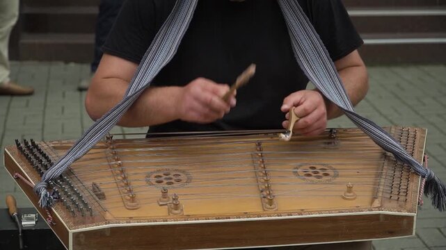 A street musician in a black T-shirt plays the ancient traditional national instrument, the dulcimer, with professional virtuosity and ease using wooden sticks, while spectators walk behind him.