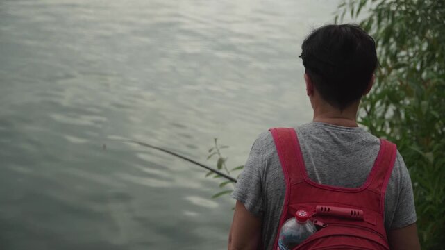 A woman with short dark hair stands shore city lake near a willow tree looks attentively and intently float fishing rod while fishing, holding large red backpack back, calm dark water