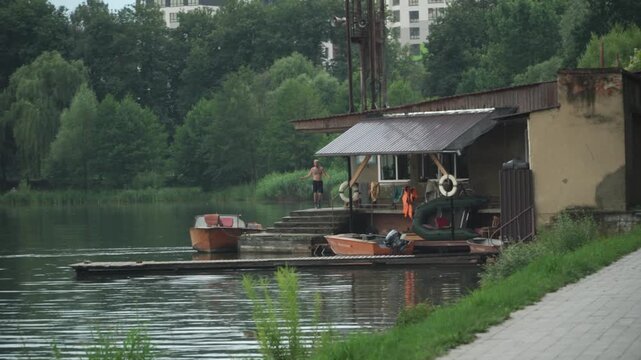 A man with athletic build black shorts jumps rope terrace house lake, small boats are moored nearby, old fishing house, lake city center, tall green trees around, cloudy summer day