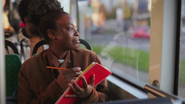 African American woman holding red notebook and pencil while sitting on bus seat and looking out window. Passenger writing and thinking about words while working or studying during commute.