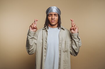 portrait of young man dressed casually is crossing his fingers, symbolizing hope and anticipation on beige background