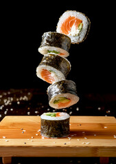 A stack of sushi rolls hovering above a wooden cutting board. The sushi rolls appear to be made with nori seaweed, rice, salmon, and avocado.