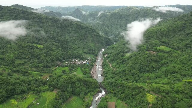 A wide aerial angle drifts to a final hold over forested mountains and a winding river. Terraced fields and a clustered village settle into the valley as mist threads through the rugged terrain.
