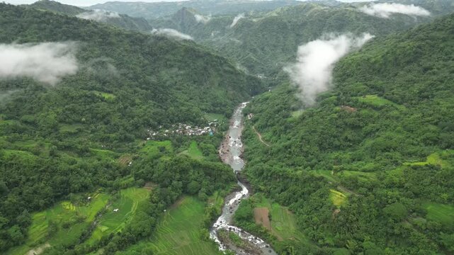A wide aerial angle drifts to a final hold over forested hills and a winding river. Mist threads through valleys as cultivated land and a small village settle into the lush green terrain.