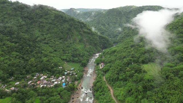 A wide aerial angle drifts to a final hold over forested mountains and a winding river. Mist threads through valleys as terraced fields and a clustered village settle into the lush terrain.
