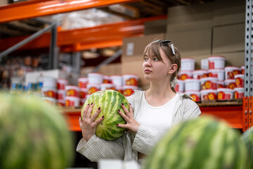 A young woman holding watermelon in budget supermarket, evaluating fruit quality during economical grocery shopping.