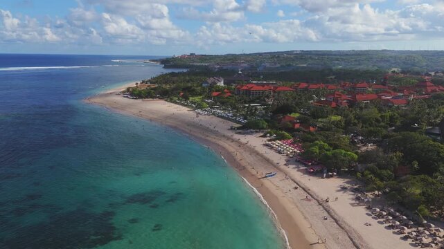 Nusa Dua resort beach in south Bali under summer sky. Clear water reveals coral patch and calm surf along the graceful arc. Family leisure and coastal walking create a relaxing holiday atmosphere