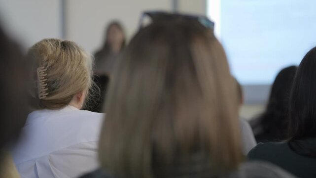 Woman with blonde hair is sitting in a classroom. She is looking at a projector screen. There are other people in the room, some of whom are also looking at the screen