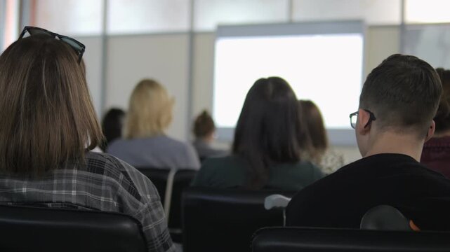 Group of people are sitting in a classroom. A woman with glasses is sitting in the front row