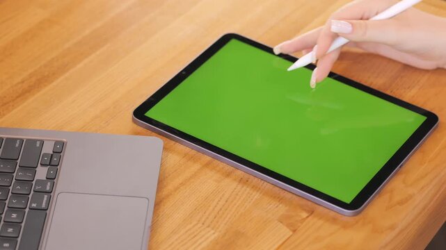 Person is using a tablet with a green screen. A laptop is also on the table. The tablet is on the right side of the laptop