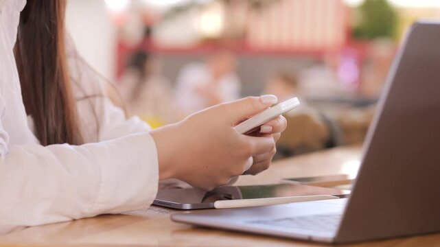 Woman is holding a cell phone in her hand. She is sitting at a table with a laptop