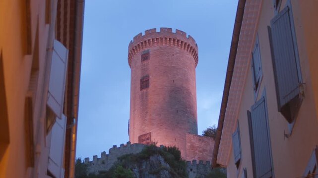 View of the illuminated medieval castle of Foix at twilight, framed by old town buildings.