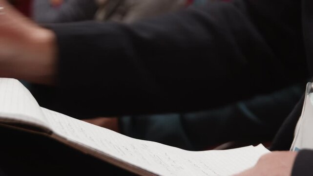 Participants are actively taking notes with a pencil on a notebook during a conference session, capturing key points and insights from the speakers.