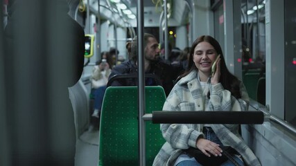 Caucasian woman with long dark hair wearing warm plaid coat sitting on bus seat holding smartphone to ear. Female smiling while talking during evening ride. Modern remote communication concept. - Powered by Adobe
