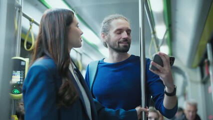 Caucasian man with beard wearing blue sweater holding smartphone showing screen to smiling woman in blue coat. Couple standing on city bus watching video together during evening ride. - Powered by Adobe
