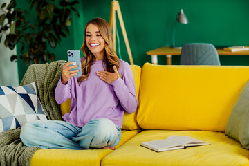 Young woman smiling on a bright sofa while holding a smartphone in a stylish interior - vibrant and...