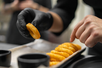 Close-up of gloved hands arranging crispy onion rings on parchment-lined tray in professional kitchen. Neat food presentation with dipping bowls in background.