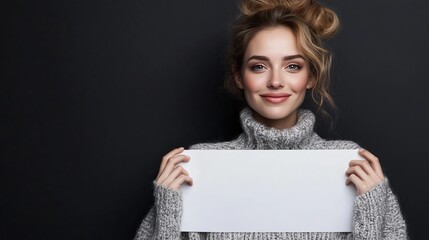 Young woman with light hair wearing a cozy gray sweater is holding a blank white sign in front of her, showcasing a friendly smile against a dark background
