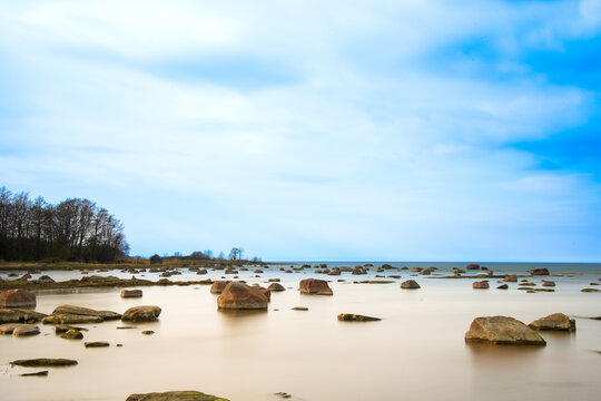 A serene shallow shoreline with scattered rocks, calm reflective water, and a soft blue sky streaked with clouds, evoking peaceful minimalism.