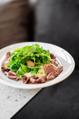 Plate of roast beef carpaccio topped with fresh arugula, creamy white sauce, and mustard dressing. Gourmet appetizer on a clean countertop, styled for culinary presentation.