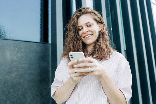 Beautiful young Caucasian wavy-haired woman uses a mobile phone while standing outdoors near a building.