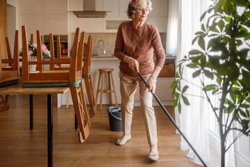 Old woman listening music through headphones and mopping parquet floor beside dining table and chairs with bucket at home