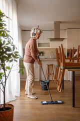 Side view of mature woman listening music over headphones and mopping parquet floor beside furniture in dining room