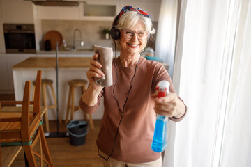 Portrait of smiling senior woman listening music over headphones holding spray bottle and napkin while cleaning house