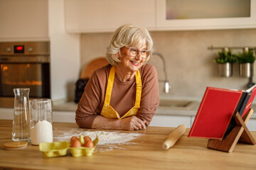 Happy senior woman in apron reading recipe book beside flour and eggs while preparing sweets for blog