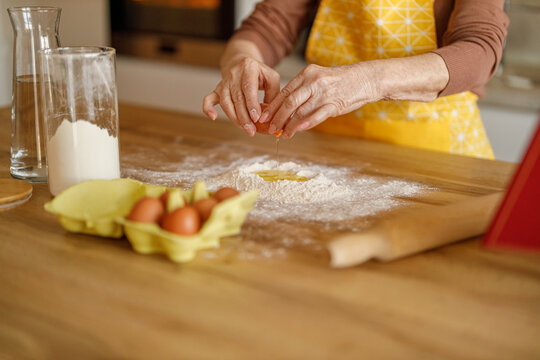 Hands of elderly woman cracking eggs into flour on kitchen counter while preparing sweets for her blog - Powered by Adobe