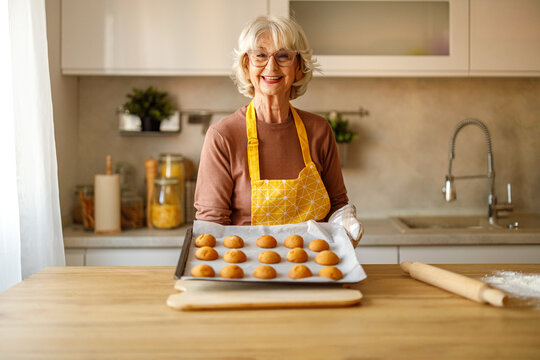 Portrait of elderly woman in apron smiling while showing tray of homemade cookies and updating her recipe blog