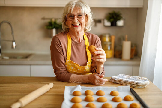 Portrait of happy senior woman smiling with tray of cookies on counter while preparing blog post at home