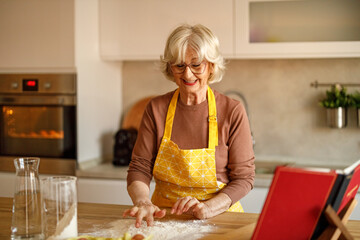 Smiling senior woman in apron mixing egg into flour on kitchen island while preparing dough for homemade cookies