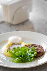 Plated breakfast with mashed potatoes, poached egg, grilled meat patty, and fresh greens on a white fluted plate. Rustic presentation on speckled countertop.