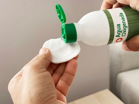 Person holding a bottle of hydrogen peroxide, pouring liquid onto a cotton pad, demonstrating personal care and hygiene practices in a clean, minimalistic environment