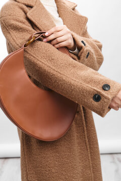 Asian woman wearing a camel coat with a leather handbag, captured in a stylish closeup. The outfit and accessories convey chic confidence and modern street style in a minimalist setting.