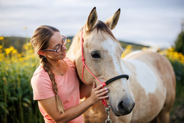 Smiling woman bonding with her horse outdoors on meadow, showing trust and emotional connection in golden evening light