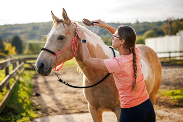 Woman grooming horse before recreational horseback riding at paddock. Female rancher using animal brush to care her paint horse