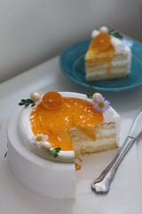 Close-up of a delicious homemade orange cake topped with whipped cream, orange glaze, and fresh tangerine slice on white background with a cake slice on a blue plate .