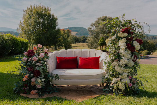 White loveseat with red cushions surrounded by pink, red, and white flowers in an outdoor garden setting, perfect for romantic occasions