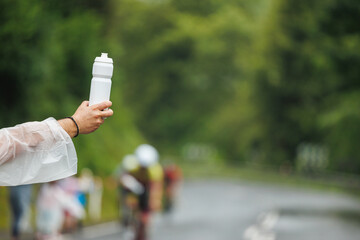 Close-up of a hand holding out a white water bottle to passing cyclists, captured in natural style with a blurred forest background, concept of hydration support