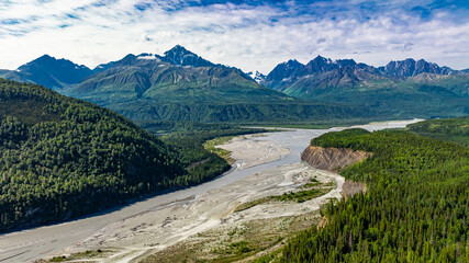 Aerial view of Alaskan river and mountain range. Wide river cutting through Alaskan forest with mountain range and cloudy sky in background.